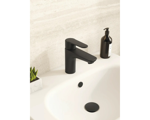 Moderne mattschwarze Waschtischarmatur auf einem weißen Waschbecken in einem hellen Badezimmer mit schwarzem Seifenspender und kleiner Pflanze.
