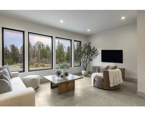 Modernes, helles Wohnzimmer mit großen Fenstern, Fliesenboden, Couchtisch aus Holz, Sofa, Sessel und einer großen Zimmerpflanze.
