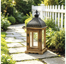 Dekorative Gartenlaterne aus Holz und Metall mit Glaseinsätzen auf einem Steinweg in einem hellen Garten mit Pflanzen und Zaun.
