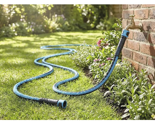 Textiler Gartenschlauch angeschlossen an einen Außenwasserhahn an einer Ziegelwand, liegend auf einer Rasenfläche im Garten.