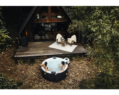 Personen entspannen in einem runden aufblasbaren Whirlpool im Außenbereich vor einem hölzernen A-Frame-Haus mit Terrasse und Lichterketten.