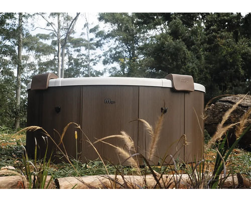 Runder MSpa Whirlpool mit Außenverkleidung in Holzoptik und zwei Kopfstützen in einer Gartenanlage.
