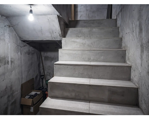 Betontreppe in einem Kellerraum mit unverputzten Wänden, beleuchtet durch eine einzelne Glühbirne.