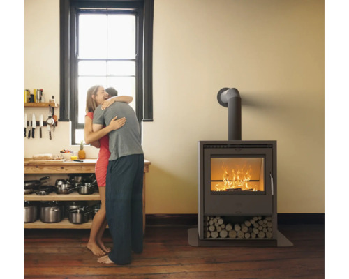 Moderner Kaminofen mit brennendem Feuer und Holzfach in einer hellen Wohnküche mit Holzfußboden und Personen im Hintergrund.