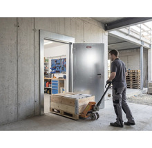 Mann zieht Hubwagen mit Holzkiste durch eine offene Abus Sicherheitstür aus Metall in ein Betongebäude mit Werkstatt im Hintergrund.