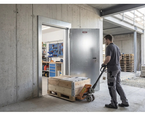 Mann zieht Hubwagen mit Holzkiste durch eine offene Abus Sicherheitstür aus Metall in ein Betongebäude mit Werkstatt im Hintergrund.