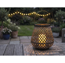 Geflochtene Laterne mit LED-Kerze auf einem Holztisch in einem Garten bei Abenddämmerung mit Lichterkette im Hintergrund.