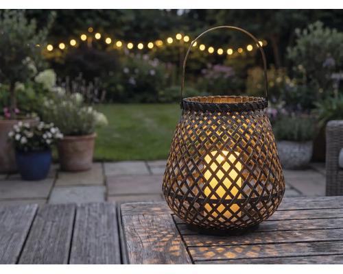 Geflochtene Laterne mit LED-Kerze auf einem Holztisch in einem Garten bei Abenddämmerung mit Lichterkette im Hintergrund.