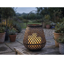 Geflochtene Gartenlaterne aus Bambus mit leuchtenden LED-Kerzen auf einem Holztisch im Garten bei Dämmerung.