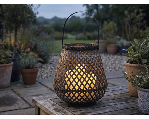 Geflochtene Gartenlaterne aus Bambus mit leuchtenden LED-Kerzen auf einem Holztisch im Garten bei Dämmerung.