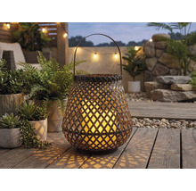 Gartenlaterne aus Flechtwerk mit Henkel und leuchtender Kerze auf einer Holzterrasse zwischen Grünpflanzen.