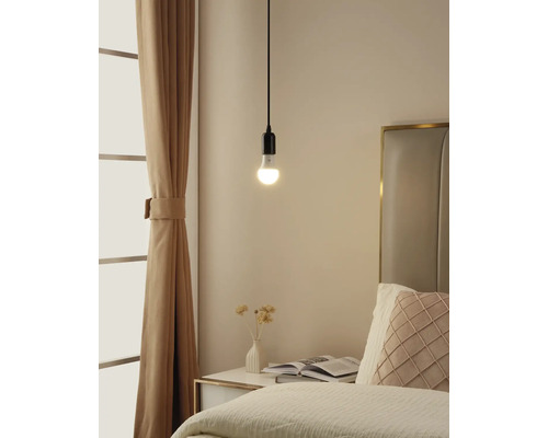 Schlichte Pendelleuchte mit brennendem Leuchtmittel über einem Nachttisch in einem hellen Schlafzimmer mit Vorhängen und Bett.