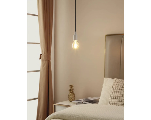 Minimalistische Pendelleuchte mit sichtbarem Leuchtmittel über einem Nachttisch in einem hellen Schlafzimmer mit Bett und Vorhang.