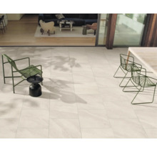 Moderne Terrasse mit hellen Bodenfliesen in Natursteinoptik, grünen Gartenmöbeln aus Metall und Übergang zum Innenraum.