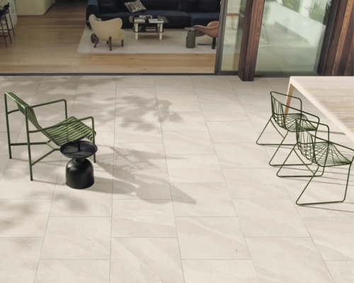 Moderne Terrasse mit hellen Bodenfliesen in Natursteinoptik, grünen Gartenmöbeln aus Metall und Übergang zum Innenraum.