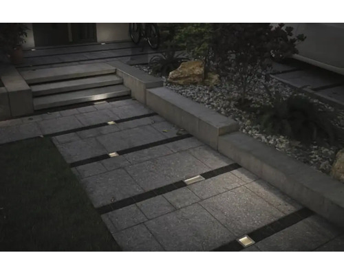 Beleuchteter Gartenweg mit rechteckigen Bodenplatten und quadratischen Bodeneinbaustrahlern in den Zwischenräumen bei Dämmerung.