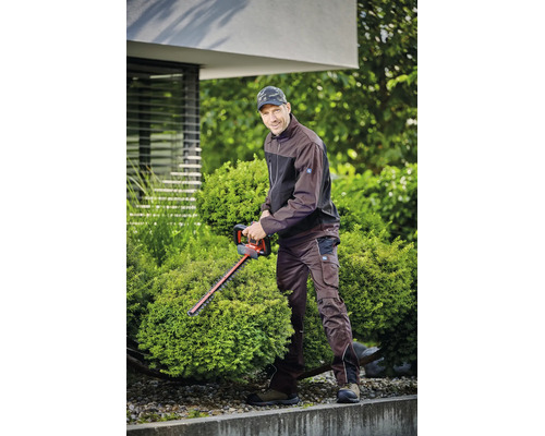 Ein Mann in brauner Arbeitsjacke und Arbeitshose schneidet einen runden Strauch mit einer Heckenschere im Garten.