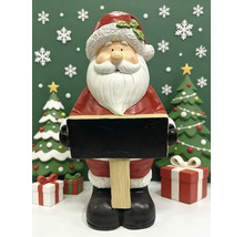 Weihnachtsmann Figur mit schwarzer Schiefertafel zum Beschriften, stehend zwischen Tannenbäumen und Geschenken.