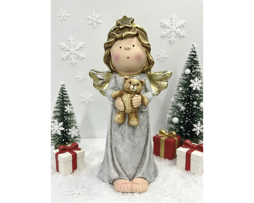 Helle Weihnachtsdekoration mit einem Engel mit goldenen Flügeln, der einen Teddybär hält, zwischen Tannenbäumen und Geschenken auf Kunstschnee.