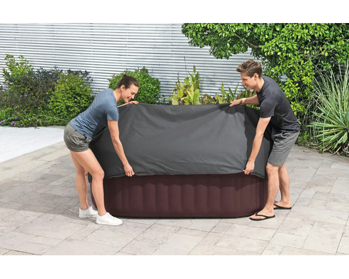 Zwei Personen legen eine graue Schutzhülle über einen aufblasbaren Whirlpool auf einer hellen Terrasse im Garten.