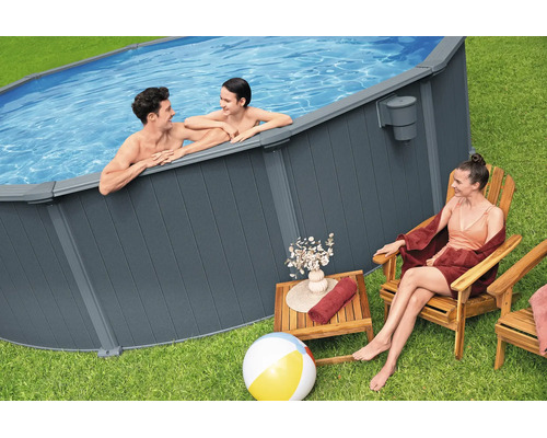 Ovaler Aufstellpool mit dunkelgrauer Lattenverkleidung im Garten. Ein Mann und ein Kind sind im Wasser, eine Frau sitzt daneben.