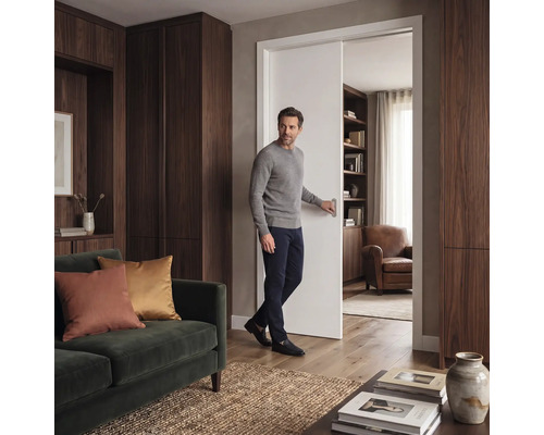 Ein Mann öffnet eine weiße Schiebetür in einem modernen Wohnzimmer mit dunklen Holzschränken, grünem Sofa und Holzfußboden.