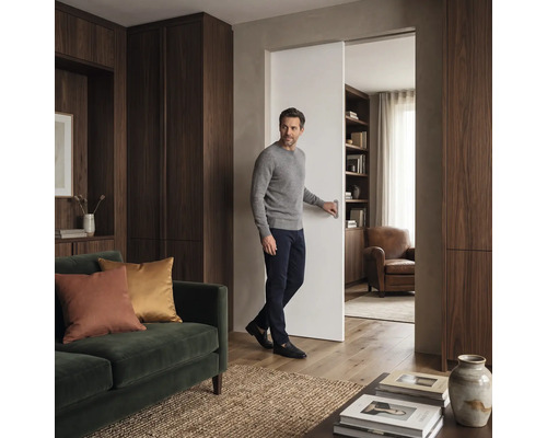 Ein Mann öffnet eine Schiebetür in einem Wohnraum mit Holzschränken, einem Sofa und angrenzendem Lesezimmer.