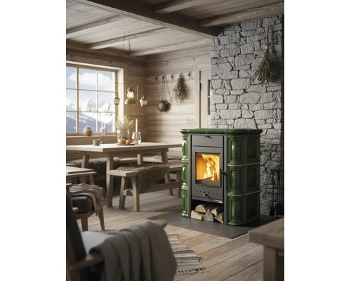 Grüner Kachelofen mit brennendem Holz in einer rustikalen Stube mit Steinwand und Holzmöbeln.