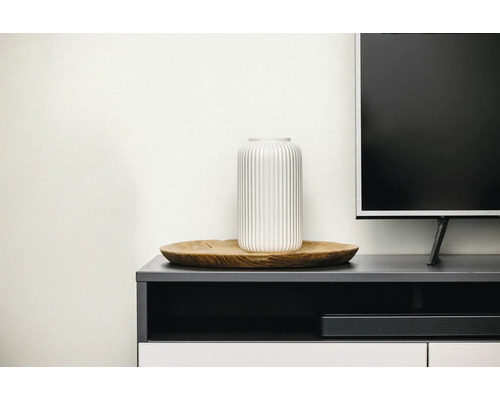 Weiße Vase mit vertikalem Rillenmuster auf einem runden Holztablett, platziert auf einem dunklen TV-Board neben einem Fernseher.