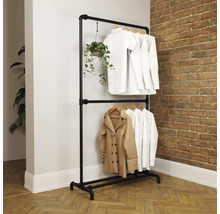Zweistöckiger Kleiderständer aus schwarzen Metallrohren im Industrial-Stil vor einer Backsteinwand, behängt mit weißer Kleidung und einer Pflanze.