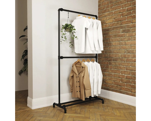 Zweistöckiger Kleiderständer aus schwarzen Metallrohren im Industrial-Stil vor einer Backsteinwand, behängt mit weißer Kleidung und einer Pflanze.