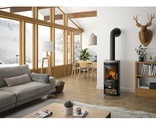 Moderner Kaminofen aus Stahl mit Sichtscheibe in einem hellen Wohnzimmer mit großen Fenstern und Blick auf eine Schneelandschaft.