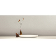 Strukturierte Wand mit rundem Podest, einer schlanken goldenen Vase mit drei dekorativen Zweigen und einem kleinen braunen geflochtenen Sessel.
