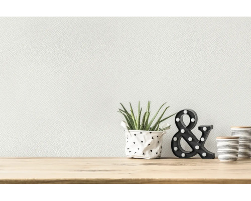 Weiße strukturierte Vliestapete an einer Wand über einem hellen Holztisch mit einer Pflanze und Dekorationselementen.