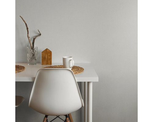 Moderner Arbeitsplatz mit weißem Schreibtisch und Stuhl vor einer grauen Wand mit Struktur. Dekoriert mit Glasvase, Holzhaus und Tasse.