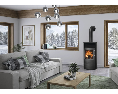 Modern eingerichtetes Wohnzimmer mit schwarzem Kaminofen Justus, grauem Sofa, Holztisch und großen Fenstern mit Blick auf eine verschneite Winterlandschaft.