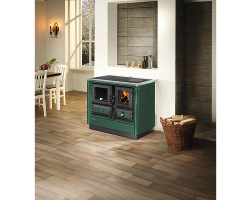 Justus Holzherd in Dunkelgrün mit brennendem Feuer in einer hellen Küche mit Holzboden und Korb mit Brennholz.