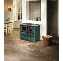 Grüner Holzherd in heller Küche mit Holzfußboden, weißem Esstisch und einem Korb mit Brennholz.