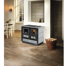 Holzherd mit Glasfront und Steinverkleidung in einer hellen Wohnküche mit Parkettboden und einem Korb voller Brennholz.