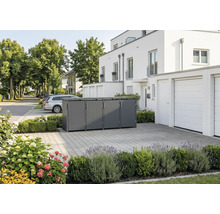 Moderne Mülltonnenbox aus Metall für vier Tonnen in einer gepflasterten Einfahrt vor einem weißen Wohnhaus mit Bepflanzung.