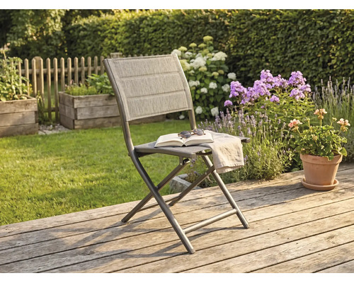 Klappbarer Gartenstuhl mit Metallgestell und Textilbespannung auf einer Holzterrasse im sonnigen Garten mit Blumen im Hintergrund.