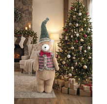 Großer Plüschbär mit Zipfelmütze und Weste neben einem geschmückten Weihnachtsbaum in einem weihnachtlich dekorierten Wohnzimmer mit Kamin.