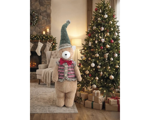 Großer Plüschbär mit Zipfelmütze und Weste neben einem geschmückten Weihnachtsbaum in einem weihnachtlich dekorierten Wohnzimmer mit Kamin.