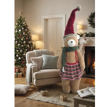 Großer Plüschbär mit Weihnachtsmütze, Schal und Rock in einem festlich dekorierten Wohnzimmer mit Weihnachtsbaum und Kamin.