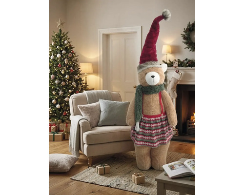 Großer Plüschbär mit Weihnachtsmütze, Schal und Rock in einem festlich dekorierten Wohnzimmer mit Weihnachtsbaum und Kamin.