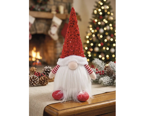 Weihnachtswichtel mit roter Paillettenhaube und weißem Kunstfellbart auf einem Tisch vor einem warmen Kamin und einem festlichen Christbaum.