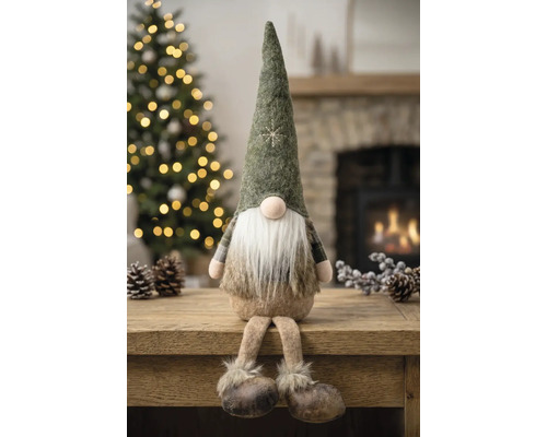 Sitzender Weihnachtsgnom mit spitzem Filzhut, langem weißen Bart und Schlenkerbeinen auf einem Holztisch in einer weihnachtlichen Umgebung.