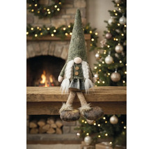 Weihnachts-Wichtel-Figur mit spitzem Hut und weißen Zöpfen, sitzend auf einem Holzbalken vor einem Kamin und Weihnachtsbaum.