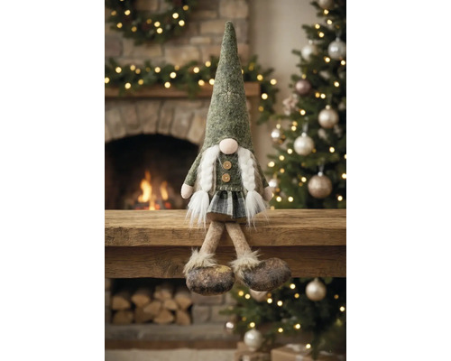 Weihnachts-Wichtel-Figur mit spitzem Hut und weißen Zöpfen, sitzend auf einem Holzbalken vor einem Kamin und Weihnachtsbaum.