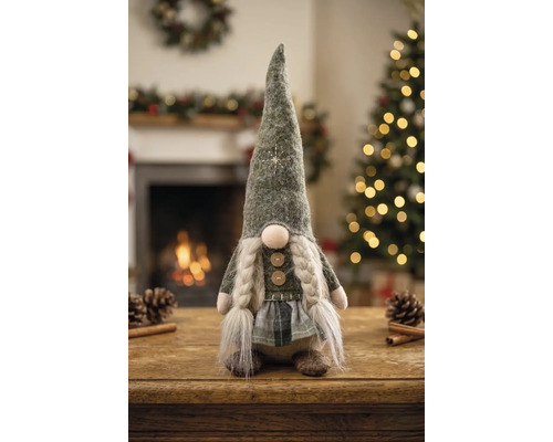 Stoff-Weihnachtsgnom mit langer Mütze, Zöpfen und Karorock auf Holztisch vor Kaminfeuer und beleuchtetem Weihnachtsbaum.